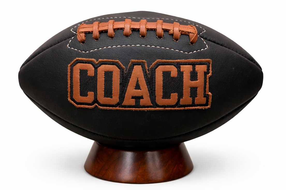 Black football-shaped pillow with 'COACH' text on a white background